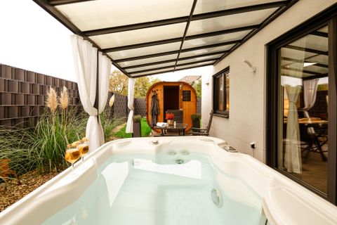 Un espace extérieur confortable avec un jacuzzi et un petit chalet en bois. Entouré de plantes vertes et d'un coin salon couvert.