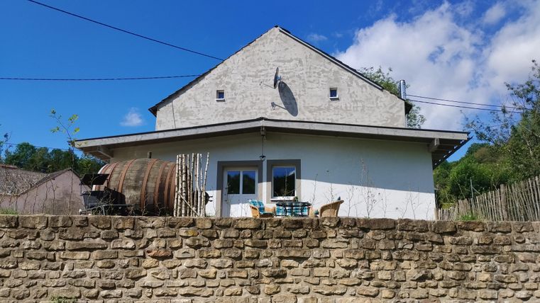 Ein charmantes Haus mit einem hellen Außenanstrich und einer Terrasse. Im Vordergrund steht eine Steinmauer und ein Holzfass.