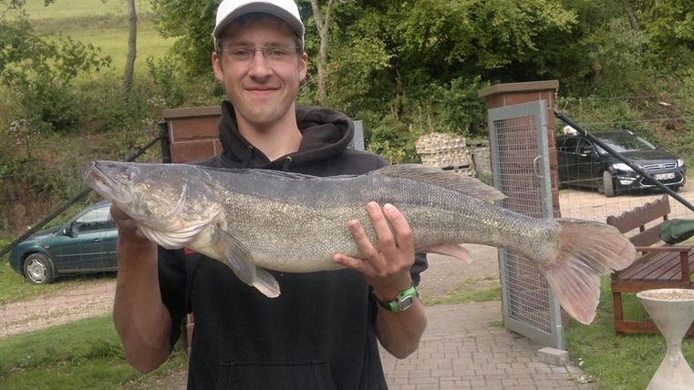 Ein junger Mann hält einen großen Fisch in den Händen. Im Hintergrund sind Bäume und Autos sichtbar.