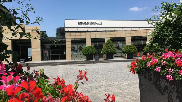 Ein modernes Gebäude mit großen Fenstern und einem klaren blauen Himmel. Im Vordergrund blühende Blumen in verschiedenen Farben.
