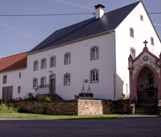 Een oude boerderij met een witte gevel en een rood dak, ernaast een kleine kapel met een kruis. De lucht is blauw en helder., © Tourist-Information Bitburger Land