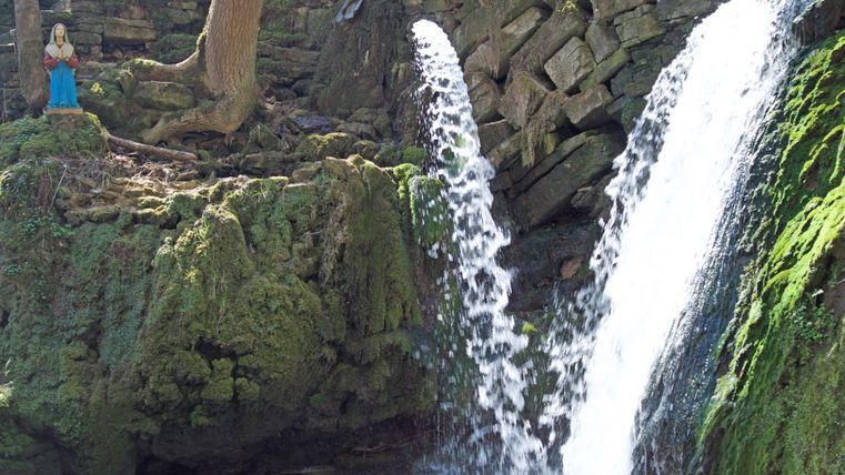 Ein kleiner Wasserfall fließt über moosbedeckte Felsen. Im Hintergrund steht eine Figur neben einem Baum.