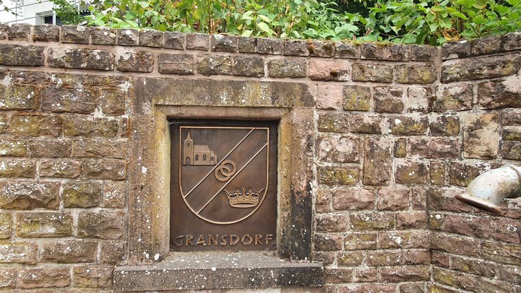 Steinwand mit Wappen von Gransdorf und einem Abflussrohr.