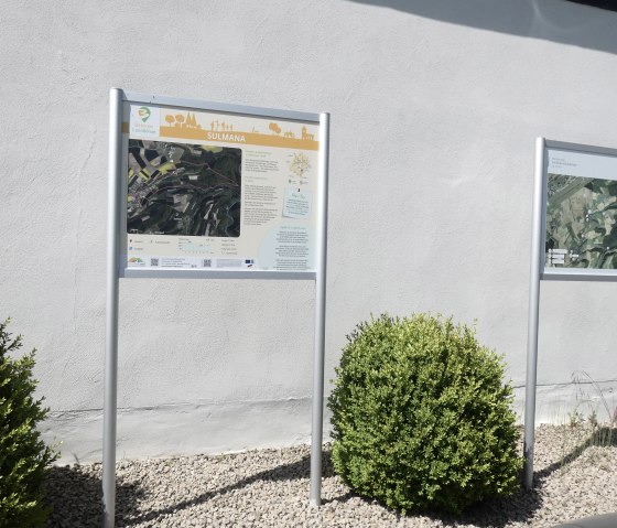 Zwei Informationstafeln mit Karten und Texten stehen vor einer weißen Wand, umgeben von grünen Büschen und Kies., © TI Bitburger Land