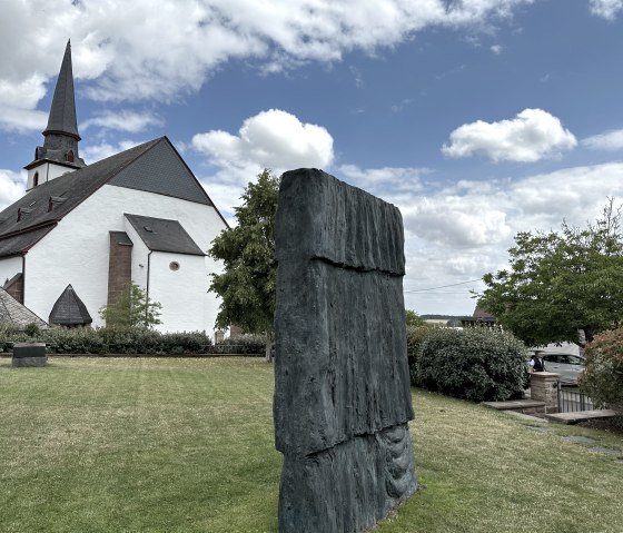 A church with a pointed tower next to a modern sculpture in the garden of the Günther Förg library in Weidingen., © Felsenland Südeifel, Anna Carina Krebs