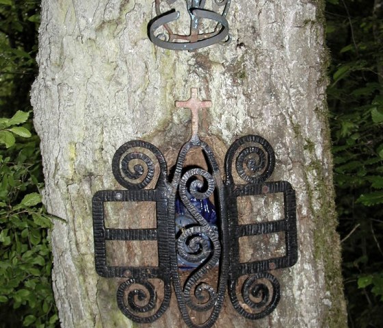Un tronc d'arbre avec un ornement métallique artistique et une petite croix. L'ornement est encastré dans l'arbre., © Conny Meier