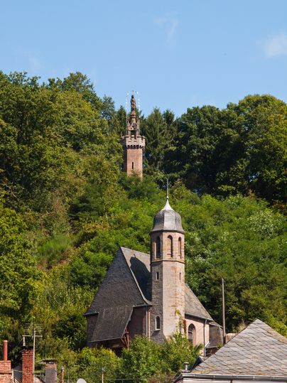 Une église avec une tour au premier plan et une colonne mariale sur une colline boisée à l'arrière-plan.