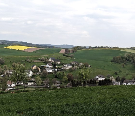 Grüne Felder und ein gelbes Rapsfeld umgeben ein kleines Dorf in Hütterscheid. Ein Spaziergänger mit Hund ist im Vordergrund zu sehen., © A. Girards