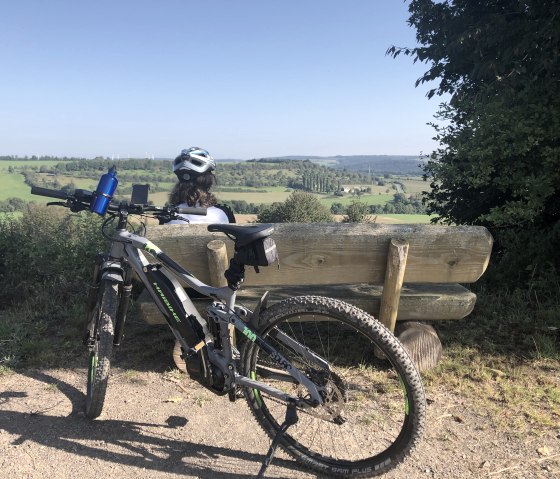 Une personne casquée est assise sur un banc et contemple un vaste paysage vert. Un vélo est posé à côté., © Tourist Information Bitburger Land