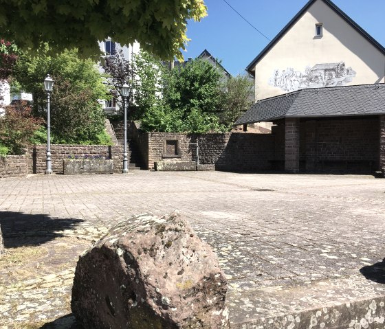 Dorfplatz in Gransdorf mit Steinmauer, Laternen und einer bemalten Hauswand. Ein Baum spendet Schatten, der Himmel ist klar und blau., © TI Bitburger Land MMayer