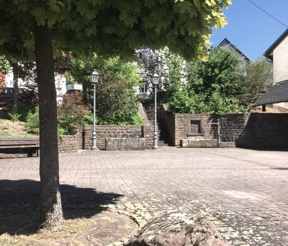 Ein gepflasterter Dorfplatz in Gransdorf mit einem großen Baum, Laternen und einem großen Stein im Vordergrund. Der Himmel ist klar und blau., © Tourist-Information Bitburger Land