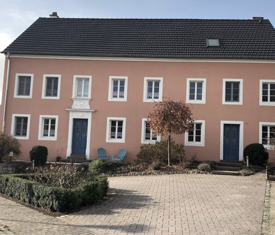 Une vieille ferme à la façade rose et aux portes bleues à Oberweis. Devant, il y a deux chaises bleues et un petit arbre dans le jardin de devant., © TI Bitburger Land - Steffi Wagner