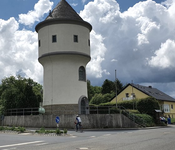 Runder Wasserturm mit spitzem Dach neben einer Straße, umgeben von Bäumen und Häusern. Ein Radfahrer fährt vorbei, der Himmel ist bewölkt.