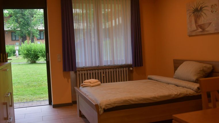 Ein einfaches Schlafzimmer mit einem Einzelbett und einem Tisch. Der Blick aus dem Fenster zeigt einen grünen Garten.