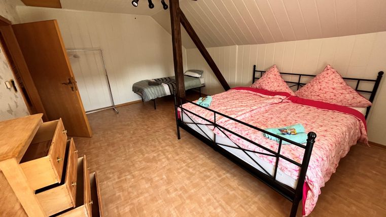 Ein gemütliches Schlafzimmer mit einem Doppelbett und rosa Bettwäsche. Neben dem Bett steht eine Kommode und es gibt einen zusätzlichen Schlafplatz in der Ecke.