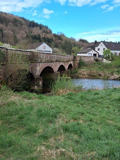 Eine historische Steinbrücke über einen kleinen Fluss, umgeben von grünen Wiesen und Bäumen. Im Hintergrund sind einige Häuser und ein klarer blauer Himmel zu sehen.