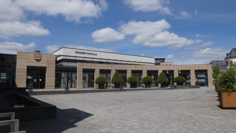 A modern building with large windows and a square in front of it. The sky is clear with some clouds.
