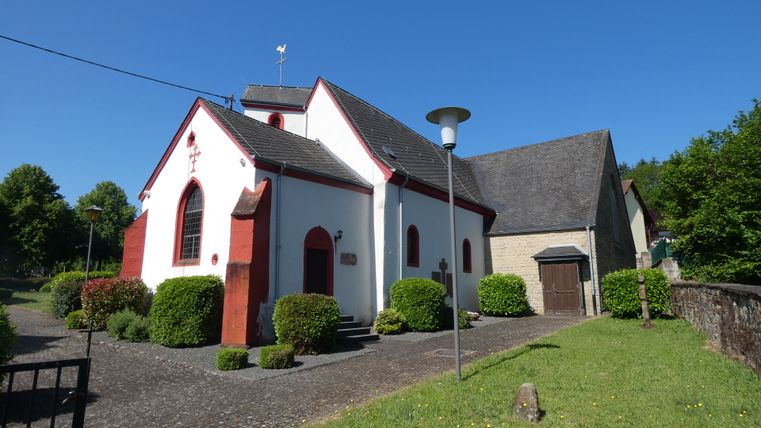 Weiße Kirche mit roten Akzenten und grauem Dach in Dahlem, umgeben von Büschen und Bäumen, bei sonnigem Wetter.