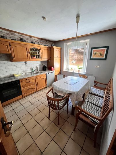 Eine helle Küche mit Holzschränken und einem großen Esstisch. Es gibt gemütliche Stühle und Fenster mit Tageslicht.