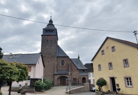 Dorfplatz mit einer Kirche aus Backstein, umgeben von bunten Häusern. Ein Baum im Vordergrund, bewölkter Himmel im Hintergrund., © TI Bitburger Land S.Wagner
