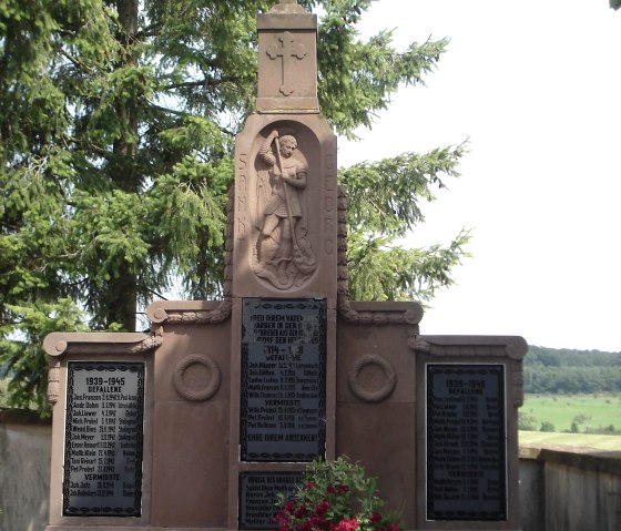 Ein Kriegerdenkmal mit St. Georg und Inschriften, flankiert von Blumen, vor einer Kulisse aus Bäumen und Landschaft., © Conny Meier