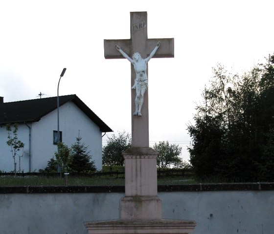 Steinkreuz mit Jesusfigur auf einem Friedhof, umgeben von Bäumen und einem Haus im Hintergrund. Inschrift: "Ich bin die Auferstehung und das Leben... 1889.", © Conny Meier
