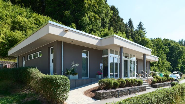Ein modernes Gebäude mit großen Fenstern und einem einladenden Eingangsbereich. Umgeben von grünen Büschen und Bäumen, strahlt es eine ruhige Atmosphäre aus.