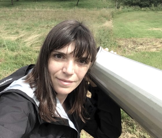 Vrouw met bruin haar houdt een grote oortrompet aan haar oor, omringd door groene weide en bomen op de achtergrond., © TI Bitburger Land M.Mayer