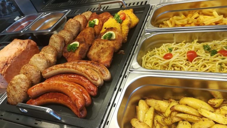Eine Auswahl von verschiedenen Fleischgerichten, darunter Würstchen und Bratlinge, ist in einer Theke präsentiert. Neben den Fleischsorten gibt es auch Nudeln und Kartoffelecken.