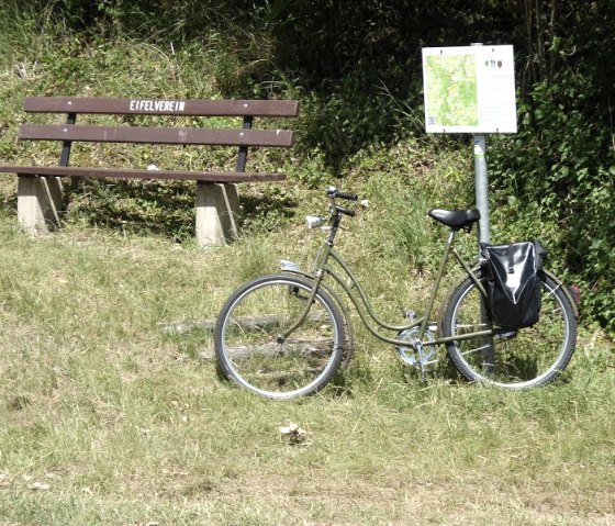 Ein Fahrrad steht neben einer Bank mit der Aufschrift 'Eifelverein' und einem Wegweiser auf einer Wiese., © Eifelverein Ortsgruppe Speicher