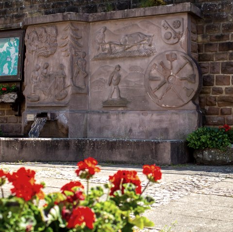 Une fontaine de village avec des reliefs artistiques en pierre, entourée de fleurs rouges. Un panneau d'information et un banc sont également visibles., © TI Bitburger Land M. Mayer