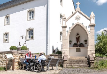 Zwei Männer, einer im Rollstuhl, sitzen auf einer Bank vor einem weißen Gebäude mit einer religiösen Statue in einer Nische. Sonniger Tag., © Naturpark Südeifel, Thomas Urbany
