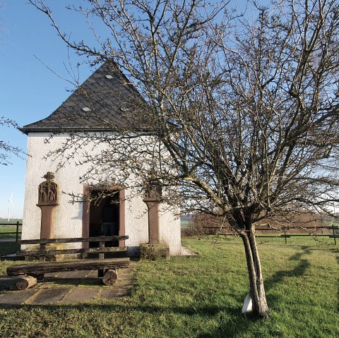 Une petite chapelle sur une prairie verte, entourée d'arbres dénudés. En arrière-plan, on aperçoit des éoliennes., © TI Bitburger Land