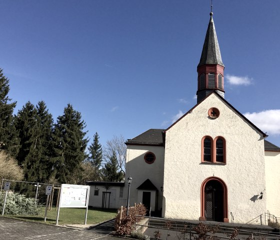A church in Wißmannsdorf with a pointed tower and red window frames against a clear blue sky., © TI Bitburger Land