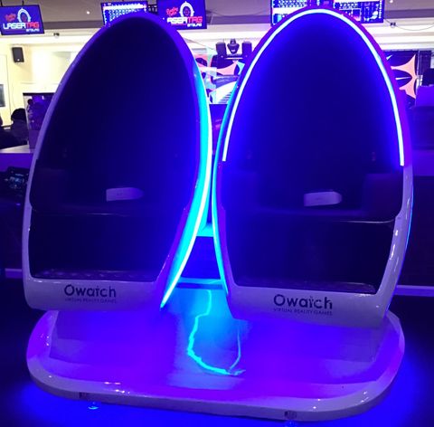 Zwei futuristische Sitze mit sanften blauen LED-Lichtern. Sie befinden sich in einem Unterhaltungskomplex und scheinen für virtuelle Realität oder immersive Erlebnisse gedacht zu sein.