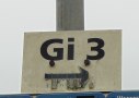 Een wit bord met de inscriptie 'Gi 3' en een pijl die naar rechts wijst, gemonteerd op een metalen paal.