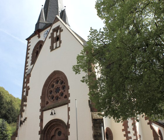 The church of St. Quirinus in Malberg with a pointed tower and a large rose window above the entrance, surrounded by green trees., © TI Bitburger Land