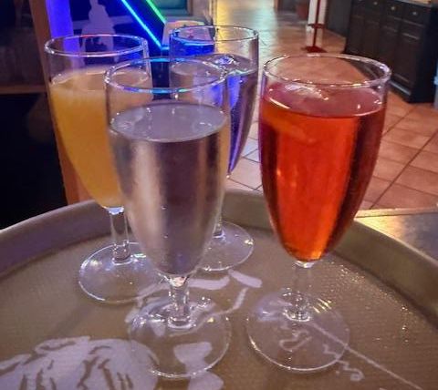 Vier Gläser mit verschiedenen Getränken auf einem Tablett. Im Hintergrund sind diffuse Lichter und eine stilvolle Inneneinrichtung sichtbar.