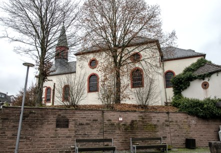 Die Kirche in Wißmannsdorf mit roten Fensterrahmen, umgeben von kahlen Bäumen und einer Steinmauer im Vordergrund., © TI Bitburger Land