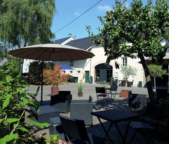 Ein Hofcafé mit Sitzgelegenheiten und Sonnenschirmen, umgeben von grünen Bäumen und Pflanzen. Im Hintergrund ein Gebäude mit der Aufschrift 'Hofcafe Messerich'., © Hofcafe Messerich