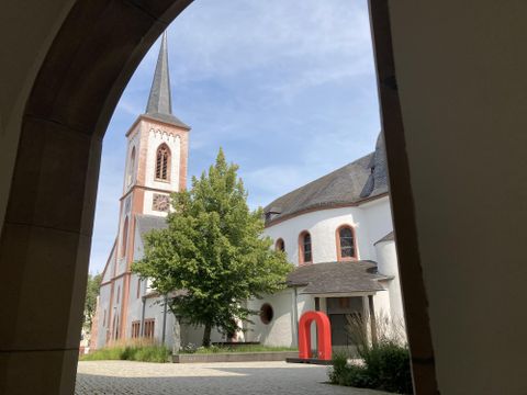 Eine Kirche mit einem hohen Turm und bunten Fenstern. Im Vordergrund sieht man einen grünen Baum und eine moderne Skulptur in Rot.