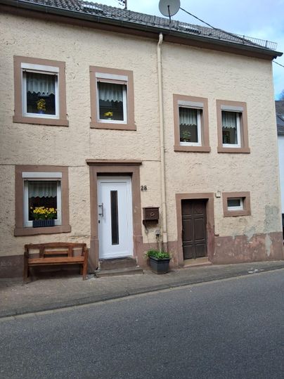 Ein gemütliches Haus an einer straße mit mehreren Fenstern und einer Eingangstür. Vor dem Haus steht eine Bank und ein Blumentopf.