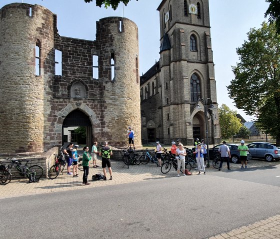 Groep fietsers voor een historische poort en een kerk, omringd door bomen en geparkeerde auto's., © Tourist-Information Bitburger Land
