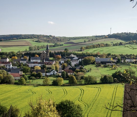 Panoramablick auf das Dorf Alsdorf mit einer Kirche im Zentrum, umgeben von grünen Feldern und Hügeln unter blauem Himmel., © Tourist-Info Bitburger Land