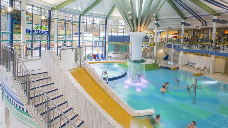 Ein modernes Schwimmbad mit einer Wasserrutsche und verschiedenen Becken. Viele Menschen genießen das Wasser und die entspannte Atmosphäre.