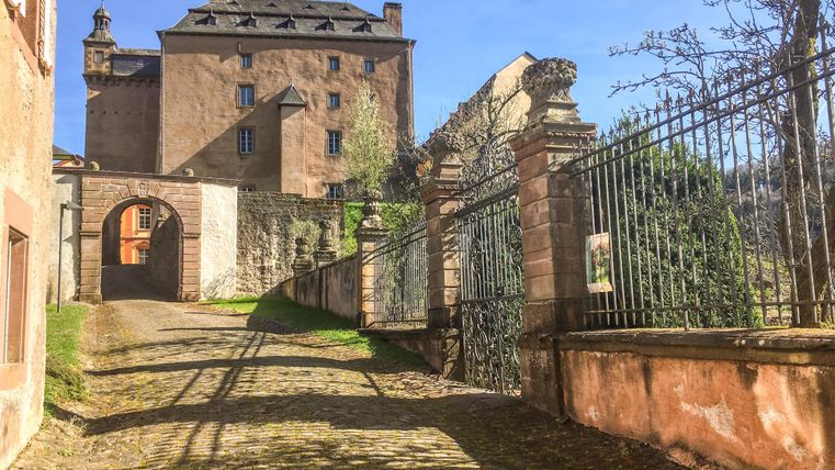 A paved path leads to an old building with a high roof. In the foreground, a wrought-iron gate and some trees can be seen.