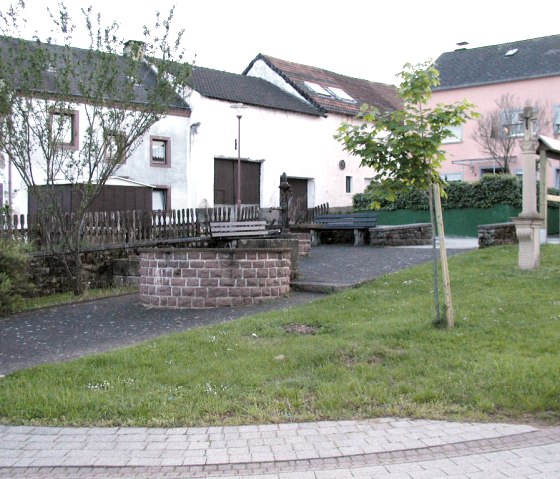 Ein kleiner Dorfplatz mit einem Brunnen, Bänken und umgeben von Häusern. Ein Baum und eine Grünfläche sind ebenfalls zu sehen., © Conny Meier
