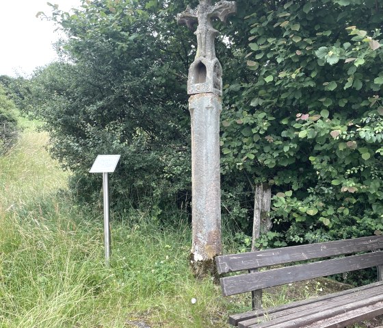A stone wayside cross stands next to a wooden bench and a sign, surrounded by green bushes and grass., © Daniel Köhler