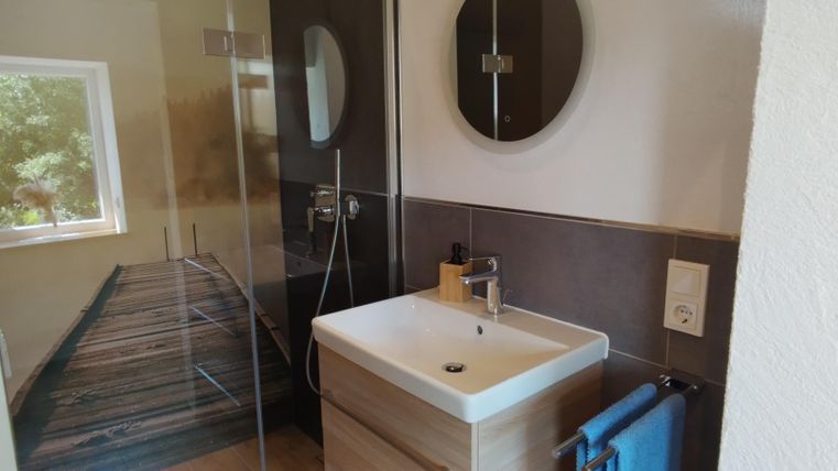 Ein modernes Badezimmer mit einer Dusche aus Glas und einem Waschbecken auf einem Holzunterbau. An der Wand hängt ein runder Spiegel und ein blauer Handtuchhalter ist sichtbar.