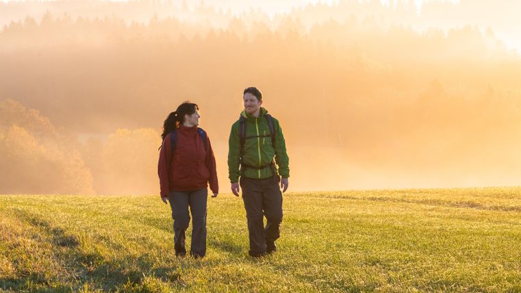 Een stel wandelt op een groen veld bij zonsopgang. De omgeving is omgeven door zachte nevelwolken en straalt een rustige sfeer uit.